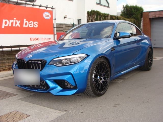 Une BMW M2 bleue devant le garage, à côté du panneau « prix bas » de la station Esso